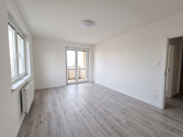 Before: empty apartment living room with grey laminate floor, white walls, a radiator under a wide window, a glazed balcony door, a doorway to the hall and a round flush ceiling light
