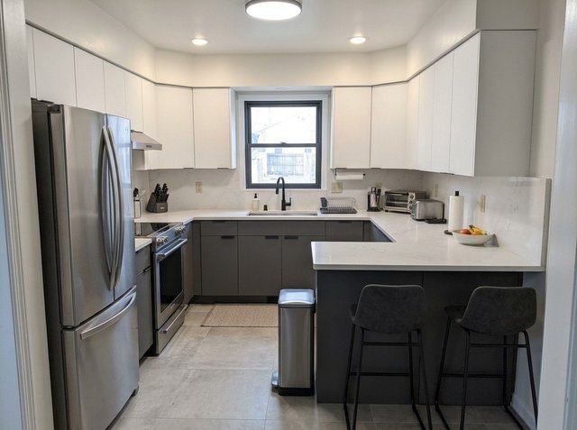 After: the same kitchen restyled as Modern, with flat white upper cabinets, gray lower cabinets, white quartz-look counters and backsplash, stainless appliances, black stools and large gray floor tiles, in the same layout