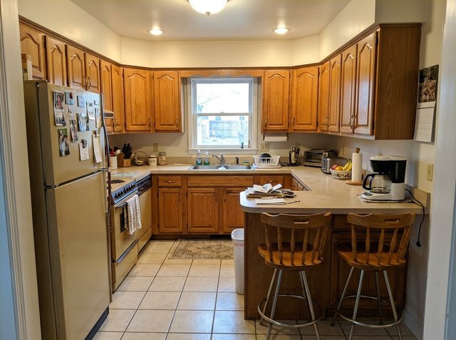 Before: a dated U-shaped kitchen with honey oak cabinets, beige laminate counters, an almond fridge and range, a peninsula with two wooden stools and cream floor tile