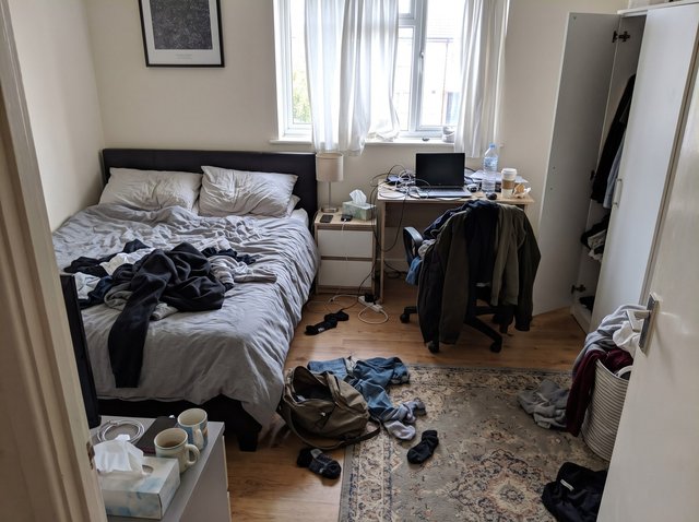 Prima: una camera da letto in disordine con vestiti e una felpa sul letto disfatto, jeans, calzini e una borsa aperta sul tappeto, giacche ammucchiate sulla sedia della scrivania, cavi aggrovigliati sulla scrivania e due tazze usate sul cassettone in primo piano