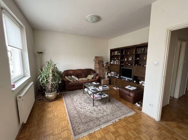 Vorher: ein bewohntes Wohnzimmer mit braunem Ledersofa, dunkler Holzwohnwand mit Fernseher, Glascouchtisch mit Zeitschriften, gemustertem Teppich, großer Pflanze und Umzugskartons auf Parkettboden