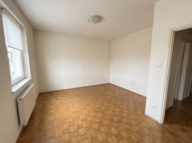 Nach Raum leeren: dasselbe Wohnzimmer mit freiem Parkettboden und weißen Wänden, Fenster mit Rollo, Heizkörper, Deckenleuchte, Türöffnung, Schalter und Steckdose bleiben erhalten