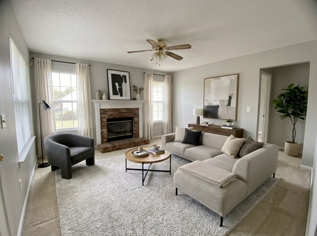 After, attempt 1: the same living room virtually staged with a light gray sectional, a gray accent chair, a round wood coffee table, a shag rug, a walnut console, a floor lamp, abstract art, a fiddle-leaf fig and linen curtains, with the carpet, fireplace and ceiling fan unchanged
