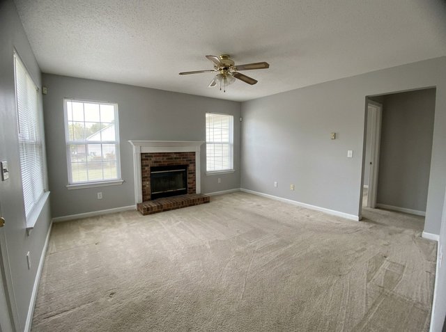 Before: an empty living room with beige carpet, light gray walls, a brick fireplace with a white mantel, two windows, a brass ceiling fan and an opening to a hallway