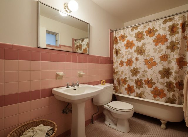 Before: a dated bathroom with pink wall tile, a pink tiled floor, a floral orange shower curtain over a clawfoot tub, a pedestal sink, a toilet and a wall mirror with a globe light