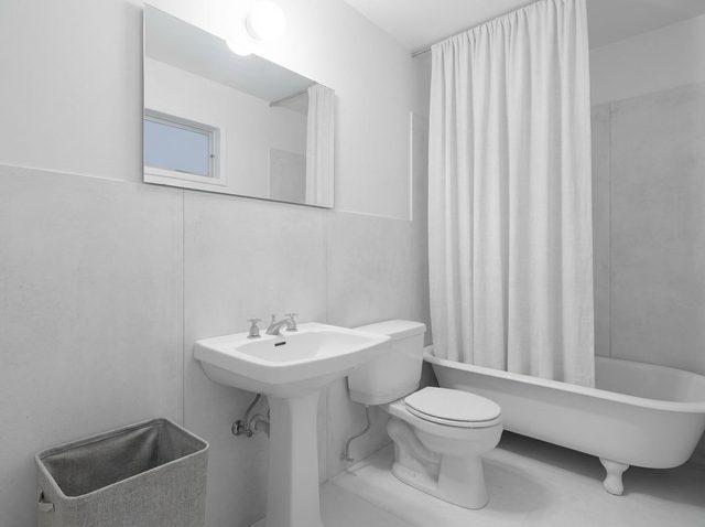 After: the same bathroom with light gray large-format wall panels, a pale floor, a white shower curtain and a gray laundry bin, keeping the pedestal sink, toilet, clawfoot tub, mirror, light and window