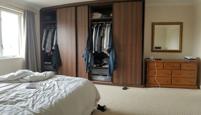 Before: bedroom with cream carpet, a walnut sliding wardrobe, a pine chest of drawers and an unmade bed